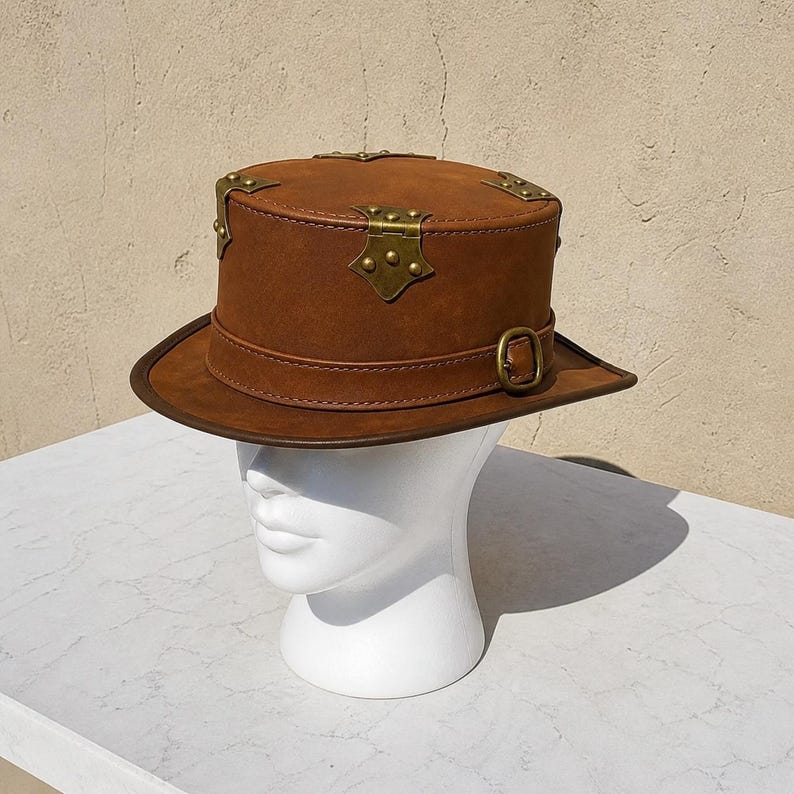 May include: A brown steampunk top hat with brass accents. The hat features a flat top, a wide brim, and a leather band with a buckle. Decorative brass corner pieces and a central clasp add to the vintage aesthetic. The hat is displayed on a white mannequin head.