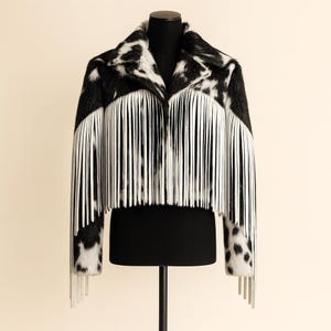 May include: A black and white cowhide-patterned jacket with long white fringe. The jacket is displayed on a black mannequin. The fringe hangs from the chest and sleeves, adding a unique texture to the garment. The jacket has a classic collar.