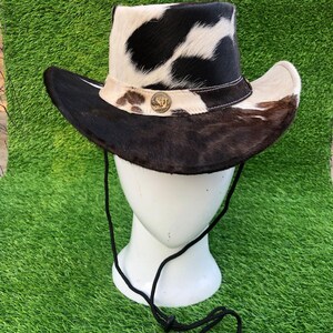May include: A cowboy hat featuring a black and white cowhide pattern on the crown and a dark brown fur brim. The hat includes a light-colored band adorned with a circular metallic emblem and a black chin cord with an adjustable toggle fastener.