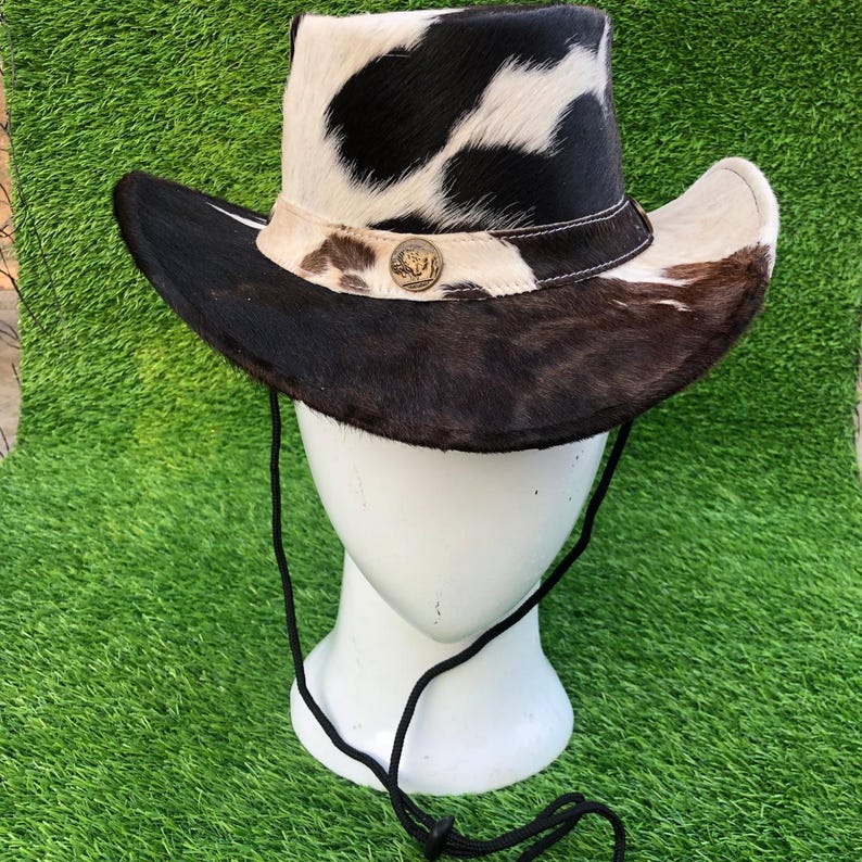 May include: A cowboy hat featuring a black and white cowhide pattern on the crown and a dark brown fur brim. The hat includes a light-colored band adorned with a circular metallic emblem and a black chin cord with an adjustable toggle fastener.