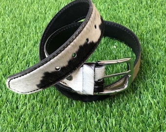 May include: A black and white cowhide belt with a silver-colored buckle. The belt is curled on a green surface, showcasing the contrasting colors and textures of the leather and metal.
