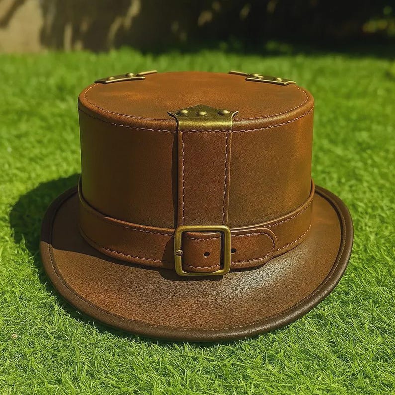May include: A brown leather top hat with a circular brim and a cylindrical crown. The hat features a brown leather strap with a brass buckle and brass accents on the crown. The hat is sitting on green grass.
