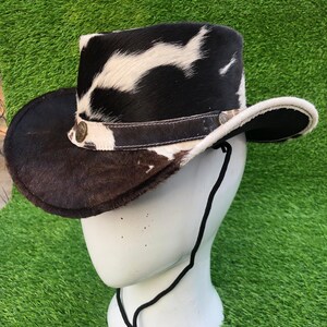 May include: A cowboy hat featuring a black and white cowhide pattern with a textured fur finish. The hat includes a dark brown leather band adorned with circular metal accents and a thin black chin cord hanging from the brim's underside.