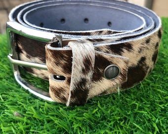 May include: A brown and tan cowhide belt with a silver buckle. The belt has a textured, spotted pattern and a gray interior. The belt is rolled up, showing the buckle and the belt loop.