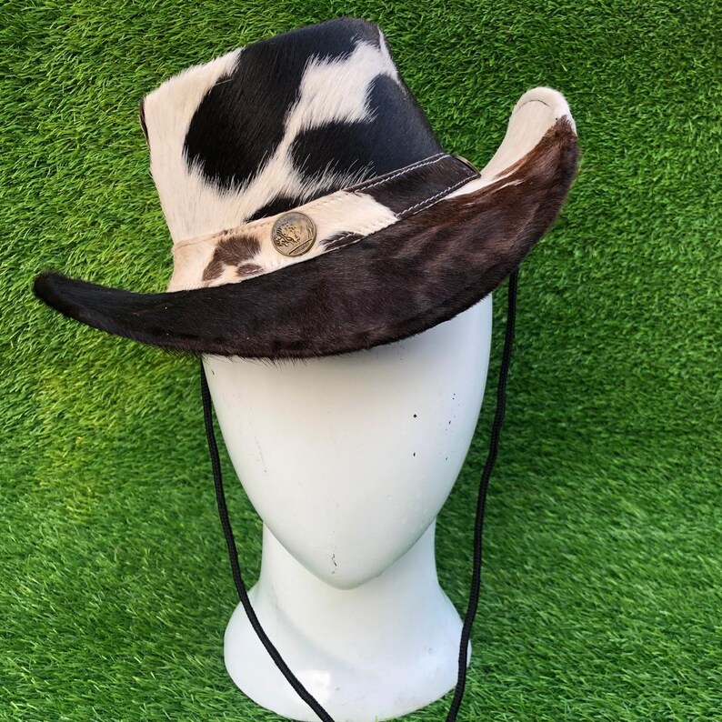 May include: A cowboy hat featuring a black and white cowhide pattern on the crown and a solid dark brown brim. The hat includes a light-colored band adorned with a circular metallic emblem and a thin black chin strap hanging from the sides.