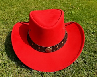 May include: A bright red cowboy hat featuring a wide brim and a pinched crown. The hat is accented with a dark brown leather band that encircles the base of the crown, decorated with small metallic studs and a central circular bronze-toned emblem.