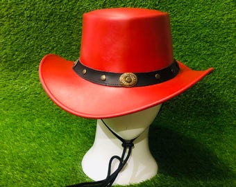 May include: A bright red cowboy hat featuring a black band adorned with metallic studs and a central oval-shaped brass-toned emblem. The hat includes a black chin cord that hangs down from the brim, providing a secure fit for the wearer.