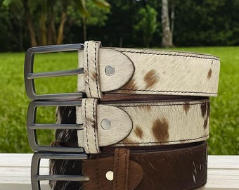 May include: Three leather belts stacked. The top two belts feature a white background with brown cowhide patterns, while the bottom belt is solid brown. Each belt has a silver buckle and a matching leather loop.