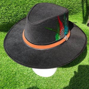 Może przedstawiać: Czarny kapelusz typu fedora z szerokim rondem i wgniecioną koroną. Kapelusz posiada jasnobrązowy skórzany pasek wokół podstawy, ozdobiony pękiem żywych zielonych, czerwonych i nakrapianych piór oraz małym okrągłym srebrnym metalowym emblematem.