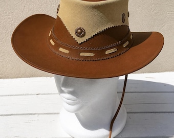 May include: A brown hat with a wide brim and a tall crown. The crown features a beige suede band with a scalloped bottom edge and three circular bronze-colored metal studs. A thin leather cord hangs from the side of the hat.