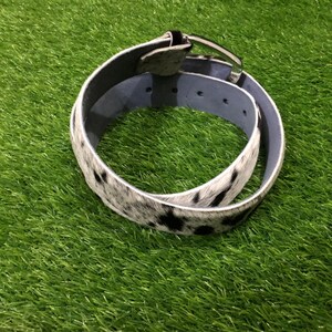 May include: A cowhide belt with a silver-tone buckle. The belt features a black and white spotted pattern on the exterior and a light blue interior. The belt is coiled on a green surface.