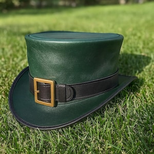 May include: A green top hat with a black band and a gold-colored buckle. The hat is made of a smooth material and sits on a bed of green grass. The hat has a wide brim and a cylindrical crown.