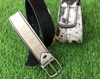 May include: Three leather belts with silver buckles are displayed on a green surface. One belt is black, another is off-white, and the third is white with brown spots. The belts are made of a cowhide material.