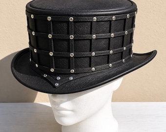 May include: A black leather top hat with a cage-like design around the crown, secured with silver-colored rivets. The hat has a wide brim and is displayed on a white mannequin head. The hat's design suggests a steampunk or gothic aesthetic.