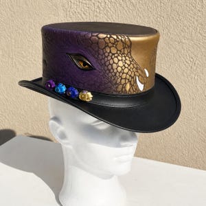 May include: A black top hat with a unique design. The hat features a purple and gold reptilian scale pattern on the crown, with an eye detail. The brim is black, and the hat is adorned with colorful jewel accents.