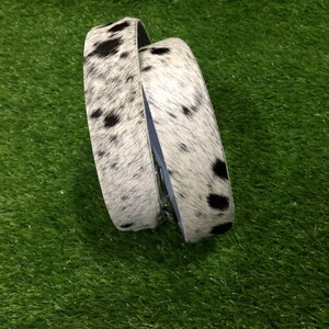 May include: A cowhide belt with a black and white spotted pattern. The belt is folded over itself, showing the interior lining. The belt is on a green surface.