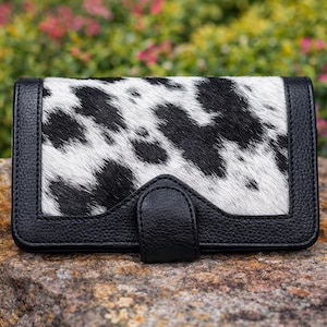 May include: A rectangular wallet with a black and white cowhide pattern on the front. The wallet has a black leather border and a black leather strap closure. The wallet is sitting on a rock.