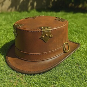 May include: A brown leather top hat with brass-colored hardware. The hat features a cylindrical crown, a wide brim, and decorative metal accents. The hat has a buckle on the side and is set against a green grass background.