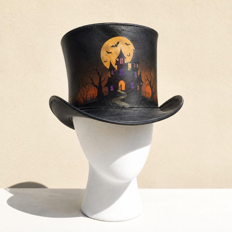 May include: A black top hat featuring a painted scene of a haunted house under a large yellow moon with flying bats. The design includes bare trees, a winding path leading to the house, and purple-lit windows against a dark, atmospheric backdrop.