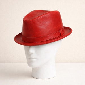 May include: A vibrant red leather fedora hat. The hat features a classic fedora design with a structured crown, a brim, and a band around the base of the crown. The hat is displayed on a white mannequin head.
