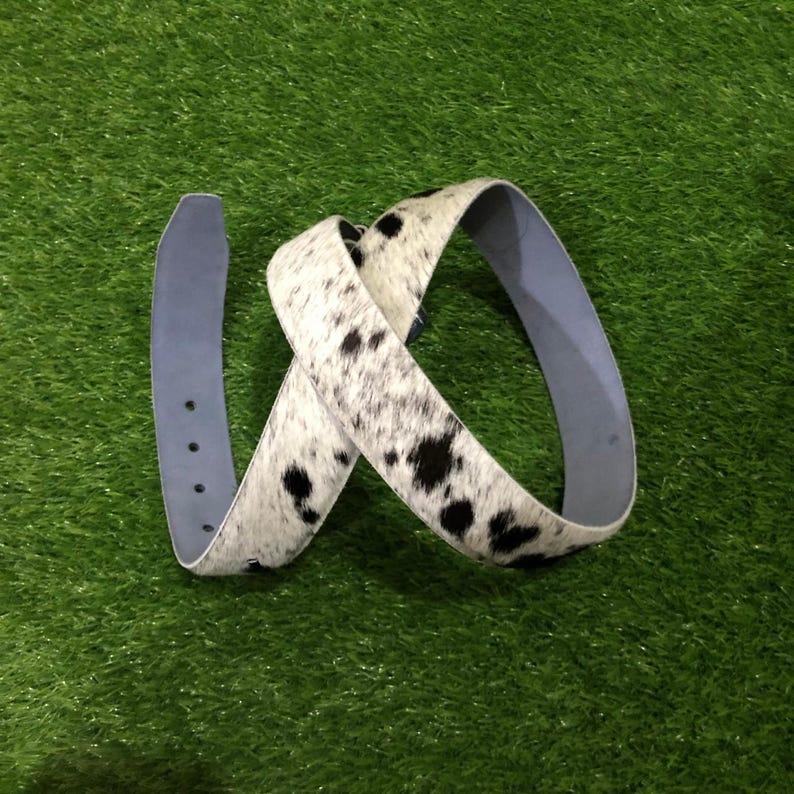 May include: A white and black cowhide belt with a light blue underside. The belt is curled and has a classic buckle design. The belt is on a green surface.