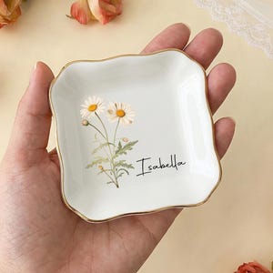May include: A small, square, pale green ceramic trinket dish with a gold edge. The dish has a floral design with two daisies and the name "Isabella" in script. The dish is held in a hand, surrounded by roses.