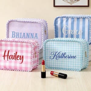 Puede incluir: Cuatro neceseres personalizados con estampados de cuadros y rayas rosas, azules y lavanda. Cada bolsa tiene un nombre bordado en un color complementario. Un lápiz labial y un frasco de perfume están en primer plano.