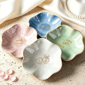 Könnte beinhalten: Vier keramik, blütenblattförmige Schalen in Pastellfarben: Rosa, Blau, Grün und Weiß. Jede Schale hat einen goldenen Rand und personalisierte Texte oder Designs. Die rosa Schale trägt den Text "woh 07.01.2026".