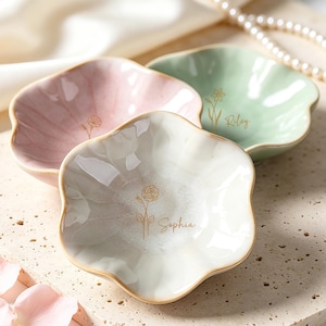 Könnte beinhalten: Drei blütenförmige Keramikschalen in Pastellfarben: Rosa, Mintgrün und Off-White. Jede Schale hat einen goldenen Rand und ein zartes Blumendesign. Die cremefarbene Schale trägt den Namen "Sophia" in goldener Schrift.