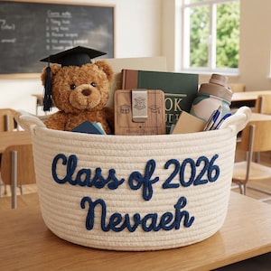 Puede incluir: Una cesta tejida color crema llena de artículos con temática de graduación, incluyendo un oso de peluche con birrete, libros, una botella de agua y un clip para billetes. La cesta lleva la inscripción "Class of 2026 Nevaeh" en letras azul marino.