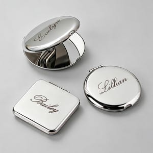 May include: Three polished silver-toned metal compact mirrors. One oval mirror is open, showing a reflective surface and the name Evelyn engraved on the lid. One square mirror is closed with the name Bailey engraved, and one round mirror is closed with the name Lillian engraved.