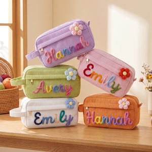 May include: Five rectangular corduroy pouches in lavender, light green, pink, white, and orange. Each pouch features a zippered closure, a side handle, and a crocheted flower accent. The names Hannah, Emily, and Avery are embroidered in colorful yarn.