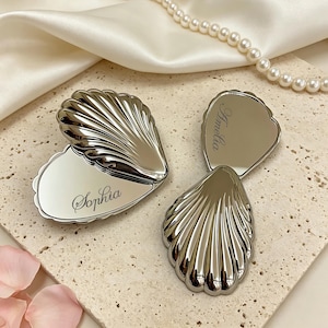 May include: Silver seashell-shaped compact mirrors with engraved names "Sophia" and "Amelia". The mirrors are open, revealing reflective surfaces. A pearl necklace and white satin fabric are in the background. The compacts are on a textured, light-coloured surface.