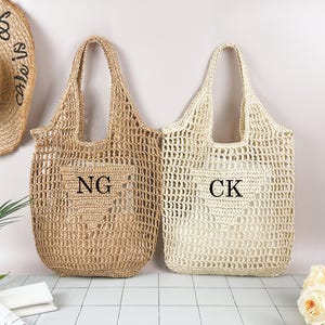 Pode incluir: Duas sacolas tote de tecido aberto, uma bege e outra creme, com alças grandes. A sacola bege tem as letras "NG" e a sacola creme tem "CK" na frente. Um chapéu de palha com as palavras "life is good" está no fundo.