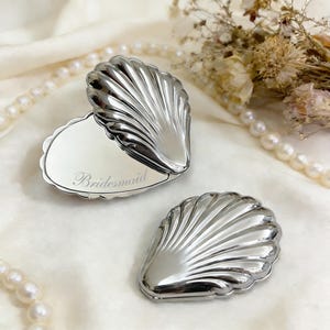 Op de afbeelding: Twee zilveren schelpvormige compacte spiegels. Eén is open en toont een spiegel met het woord "Bridesmaid" gegraveerd. De andere spiegel is gesloten. De spiegels liggen op een witte ondergrond, met parels en droogbloemen op de achtergrond.