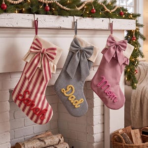 May include: Three Christmas stockings hanging from a mantel. The first is red and white striped with a red bow and the name Sarah. The second is black and white gingham with a matching bow and the name Dad. The third is solid mauve with a mauve bow and the name Mom.