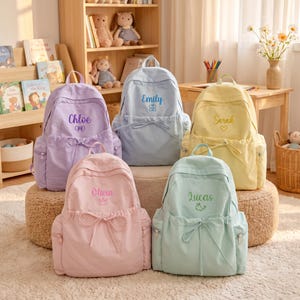 May include: Five pastel-coloured backpacks featuring front drawstring pockets and embroidered names. The purple bag says Chloe, blue says Emily, yellow says Sarah, pink says Olivia, and mint green says Lucas. Each bag has a small icon below the name.