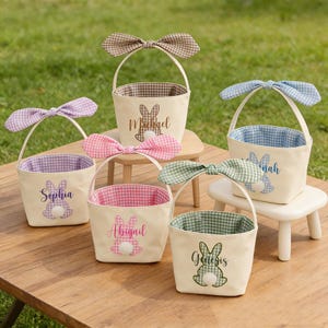 May include: Six cream-colored Easter baskets with bunny ear handles and gingham linings. Each basket has a different colored bow and is personalized with a name and bunny graphic. The baskets are arranged on a wooden table.
