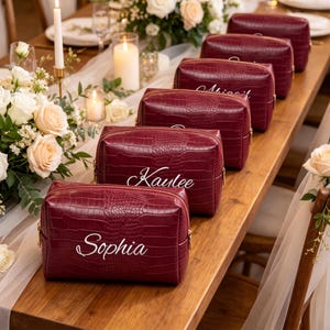 May include: A series of rectangular burgundy cosmetic bags with a crocodile skin texture, each featuring a white cursive name printed on the front. The bags are arranged in a line on a wooden surface, with visible names including Sophia, Kaulee, and Abigail.