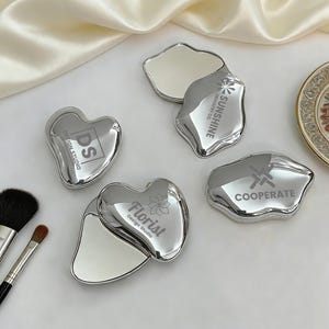 May include: A collection of silver-colored compact mirrors in various shapes, including heart and irregular forms. Each mirror has a reflective surface and is engraved with logos and text such as "Design Studio", "Sunshine Industry Co.", and "Florist Design Studio".