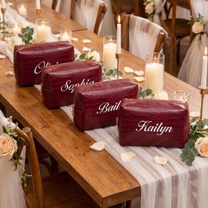 May include: Four rectangular burgundy cosmetic bags with a crocodile-embossed texture and gold-tone zips. Each bag features a white embroidered name in a cursive script, specifically the names Olivia, Sophia, Bailey, and Kailyn, displayed clearly.