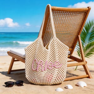 Op de afbeelding: Een cr&egrave;mekleurige gehaakte tote bag met een open geweven mesh-ontwerp en twee lange schouderbanden. De voorkant van de tas is voorzien van de naam Chloe in verhoogde, zachtroze cursieve letters. De tas is gemaakt van gestructureerd garen.