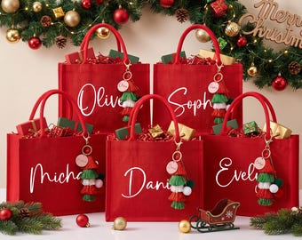 May include: Five red tote bags with red handles, each personalized with a white name in script font. Each bag features a decorative keychain with a round wooden tag and a multi-colored tassel in red, green, and white, filled with gift boxes.