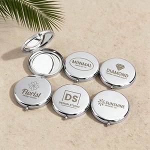 May include: A collection of six round, silver-toned metal compact mirrors. One mirror is open, revealing a reflective surface. The other five are closed, each featuring a unique logo: Minimal Industry Co., Diamond, Florist Design Studio, DS Design Studio, and Sunshine Industry Co.