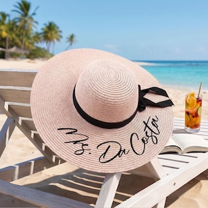 May include: A light pink sun hat with a wide brim, a black ribbon, and the text "Mrs. Da Costa" in black script. The hat is on a white beach chair near a tropical beach with a cocktail and a book.