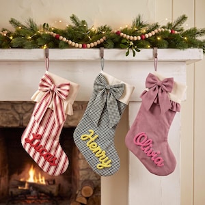May include: Three hanging Christmas stockings with cream-coloured cuffs and bows. The first is red and white striped with the name Lucas in red. The second is black and white gingham with the name Henry in yellow. The third is mauve with the name Olivia in pink.