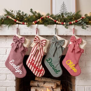 May include: Four personalised Christmas stockings hanging in a row. The first is dusty rose with white text, the second is red and white striped with red text, the third is green gingham with green text, and the fourth is red gingham with yellow text.