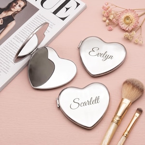 Puede incluir: Tres espejos de bolsillo plateados con forma de corazón. Un espejo está abierto y muestra una superficie reflectante. Dos espejos cerrados presentan los nombres grabados Evelyn y Scarlett en letra cursiva. Los espejos tienen un acabado metálico pulido.