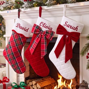 May include: Three Christmas stockings hanging in a row. The first is red plaid with a white cuff embroidered with the name David. The second is solid red with a quilted texture, a plaid bow, and a white cuff embroidered with the name Olivia. The third is white with a quilted texture, a large red bow, and a white cuff embroidered with the name Sarah.