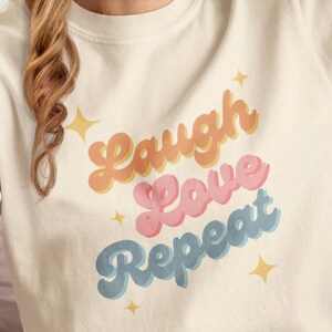 May include: A white t-shirt with a retro-style graphic that says "Laugh Love Repeat" in colorful, layered lettering. The graphic is surrounded by small gold stars.